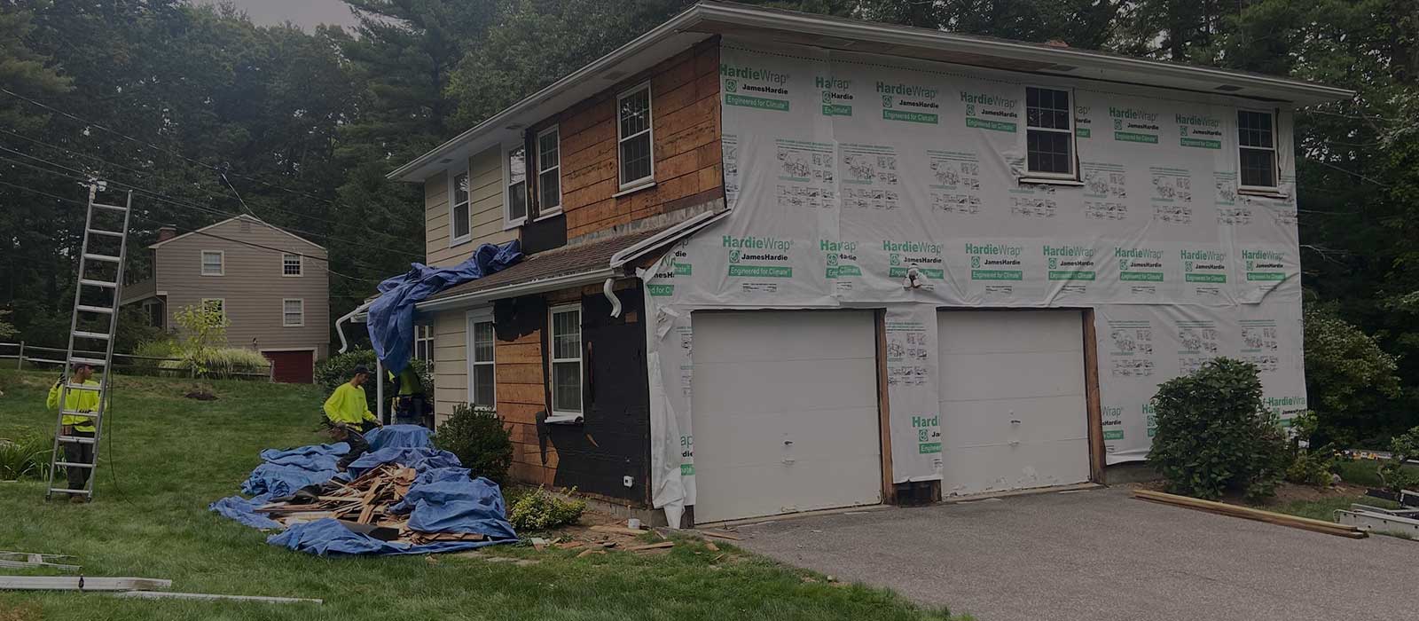 A two-story home with the siding removed, covered in HardieWrap insulation as workers haul away debris.